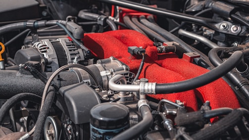 A detailed view of a car engine, featuring various components such as pipes, hoses, and wires. The engine has a prominent red part, likely a manifold cover, standing out against the metallic and black hues of the surrounding components.