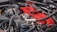 A detailed view of a car engine, featuring various components such as pipes, hoses, and wires. The engine has a prominent red part, likely a manifold cover, standing out against the metallic and black hues of the surrounding components.