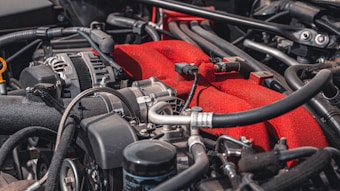 A detailed view of a car engine, featuring various components such as pipes, hoses, and wires. The engine has a prominent red part, likely a manifold cover, standing out against the metallic and black hues of the surrounding components.