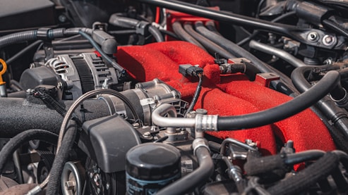 A detailed view of a car engine, featuring various components such as pipes, hoses, and wires. The engine has a prominent red part, likely a manifold cover, standing out against the metallic and black hues of the surrounding components.