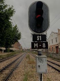 A railroad track stretches into the distance with a traffic signal displaying a red light. The scene is flanked by residential buildings and a tree on the left. Overhead wires span the tracks, and there is a weathered control box with graffiti at the base of the signal.