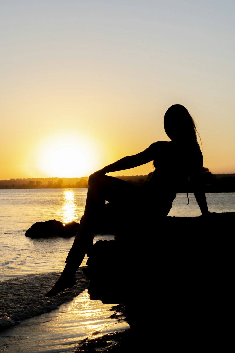 日没時の水域近くの岩の上に座っている女性のシルエット