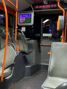 The interior of a public bus with empty seats and orange handrails. A digital screen displaying information is mounted above the seating area. The digital strip near the ceiling shows the time and date. The overall setting is dimly lit.