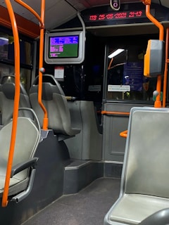 The interior of a public bus with empty seats and orange handrails. A digital screen displaying information is mounted above the seating area. The digital strip near the ceiling shows the time and date. The overall setting is dimly lit.
