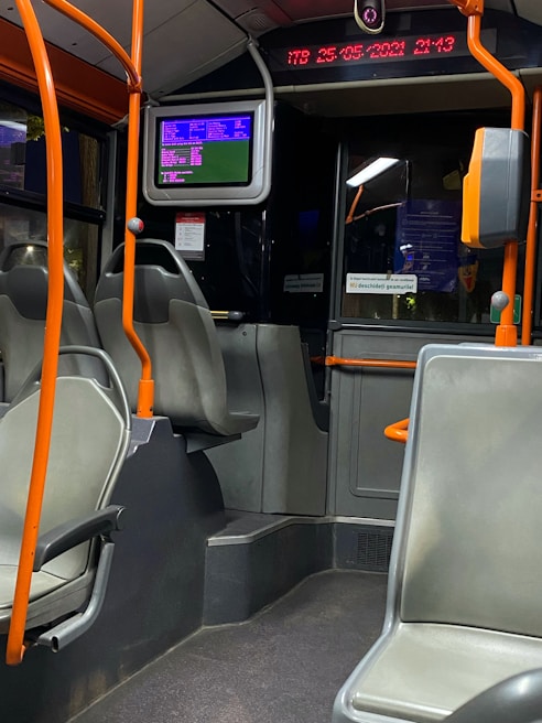 The interior of a public bus with empty seats and orange handrails. A digital screen displaying information is mounted above the seating area. The digital strip near the ceiling shows the time and date. The overall setting is dimly lit.