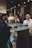 A bustling bar scene with a bartender preparing drinks, surrounded by guests engaged in conversation. The setting is dimly lit with a warm ambiance, featuring decorative lighting. A bouquet of white flowers adds a touch of elegance to the countertop, and various bottles are visible near the bar area.