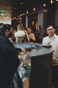 A bustling bar scene with a bartender preparing drinks, surrounded by guests engaged in conversation. The setting is dimly lit with a warm ambiance, featuring decorative lighting. A bouquet of white flowers adds a touch of elegance to the countertop, and various bottles are visible near the bar area.
