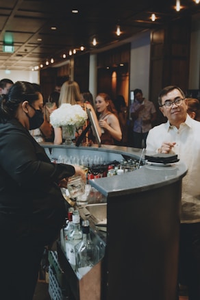 A bustling bar scene with a bartender preparing drinks, surrounded by guests engaged in conversation. The setting is dimly lit with a warm ambiance, featuring decorative lighting. A bouquet of white flowers adds a touch of elegance to the countertop, and various bottles are visible near the bar area.