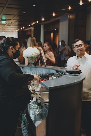A bustling bar scene with a bartender preparing drinks, surrounded by guests engaged in conversation. The setting is dimly lit with a warm ambiance, featuring decorative lighting. A bouquet of white flowers adds a touch of elegance to the countertop, and various bottles are visible near the bar area.