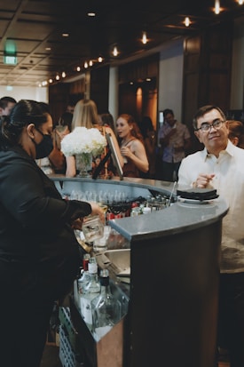 A bustling bar scene with a bartender preparing drinks, surrounded by guests engaged in conversation. The setting is dimly lit with a warm ambiance, featuring decorative lighting. A bouquet of white flowers adds a touch of elegance to the countertop, and various bottles are visible near the bar area.