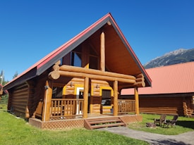 A wooden log cabin with a steep red roof and a front porch with railings. The structure is made of large logs, and it sits on a green lawn. Nearby, there are chairs around a small fire pit. In the background, mountains are visible under a clear blue sky.