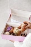 A hand is holding an open pastel pink box containing an assortment of decorated pastries. Each pastry is filled with different colored creams and toppings, including purple, white, green, and chocolate with various garnishes.