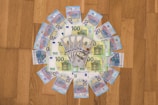 A circular arrangement of assorted Euro banknotes and a few US dollar bills are placed on a wooden surface. The Euro notes are visually dominant, with 100 and 20 denominations prominently visible.