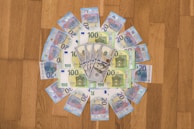 A circular arrangement of assorted Euro banknotes and a few US dollar bills are placed on a wooden surface. The Euro notes are visually dominant, with 100 and 20 denominations prominently visible.