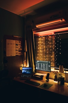 A cozy workspace setup with warm lighting includes a desk with a laptop, a desktop monitor displaying a music playlist, a keyboard, and a cup. The room features hanging string lights near a window, casting a soft glow, and is decorated with a plant, a lamp, and a calendar on the wall.