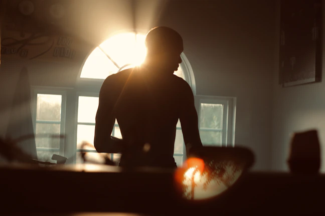 A serene portrait of a mature mother in a soft, elegant fitness outfit standing by a sunlit window.