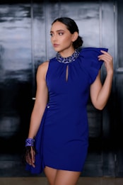 A woman in a glamorous, blue one-shoulder dress adorned with a beaded collar stands in front of a dark, textured background. Her hair is styled in an elegant updo, and she wears a matching blue bracelet with feather details.