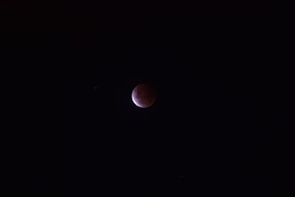 A dark sky with a partially illuminated circular object, suggesting a lunar eclipse. The moon displays a deep reddish color on one side while the rest remains shrouded in shadow. A few faint stars are scattered in the vast darkness around it.