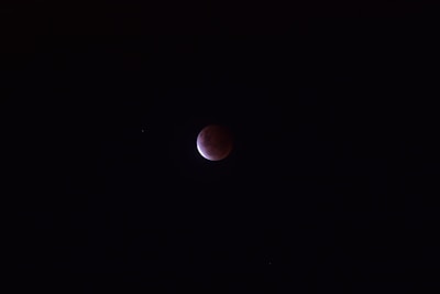A dark sky with a partially illuminated circular object, suggesting a lunar eclipse. The moon displays a deep reddish color on one side while the rest remains shrouded in shadow. A few faint stars are scattered in the vast darkness around it.