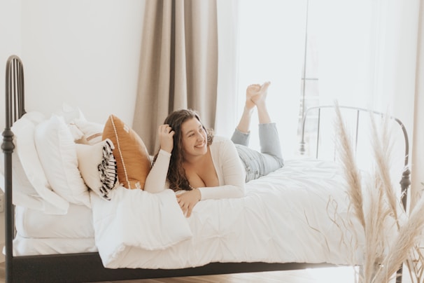 A person is lying on a bed covered with white sheets, surrounded by pillows in light shades. The room is softly lit by natural light coming from a window with sheer curtains. Decorative grass stalks can be seen in the foreground, complementing the cozy and relaxed atmosphere.