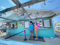A small food stand with a turquoise exterior has several people ordering and waiting in line. The setup includes colorful signage, menus displayed in frames, and an 'Open' sign. A wooden trellis covered with fishing nets and decor like a model of a shark is seen overhead. The setting appears casual and coastal.