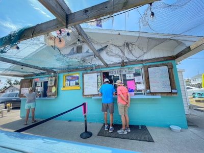 A small food stand with a turquoise exterior has several people ordering and waiting in line. The setup includes colorful signage, menus displayed in frames, and an 'Open' sign. A wooden trellis covered with fishing nets and decor like a model of a shark is seen overhead. The setting appears casual and coastal.