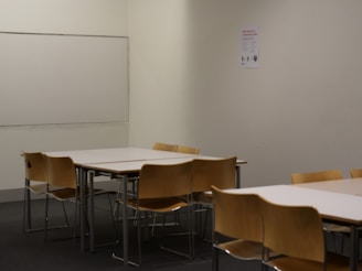Several wooden chairs are arranged around two rectangular tables in a room with a plain light-colored wall. A whiteboard is mounted on the wall alongside a small poster.