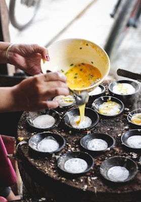 A person is pouring a yellow batter from a bowl into small round black pans on a cooking surface. The batter appears to contain green herbs, and the steam indicates that these are being cooked. The image also hints at a street or an outdoor setting due to the blurred background.