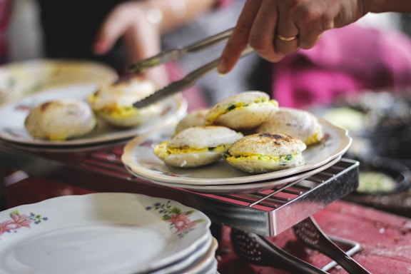 A person is using tongs to serve a plate of steaming hot idlis, an Indian dish. The idlis are neatly arranged on a floral plate placed on a wire rack over a red tablecloth. The surrounding environment suggests a casual dining setting.