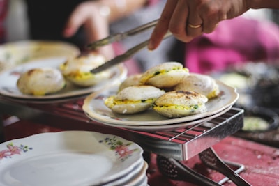 A person is using tongs to serve a plate of steaming hot idlis, an Indian dish. The idlis are neatly arranged on a floral plate placed on a wire rack over a red tablecloth. The surrounding environment suggests a casual dining setting.