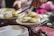 A person is using tongs to serve a plate of steaming hot idlis, an Indian dish. The idlis are neatly arranged on a floral plate placed on a wire rack over a red tablecloth. The surrounding environment suggests a casual dining setting.