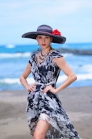 A woman stands on a beach wearing a stylish black and white patterned dress and a large wide-brimmed hat decorated with a red flower. The background features a serene ocean view with gentle waves and a blurred horizon.