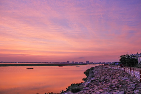 A serene lake is bathed in the warm hues of a sunset, with vibrant oranges, pinks, and purples spreading across the sky. The calm water reflects the sky's colors, creating a mirror-like effect. A paved pathway lined with a railing runs alongside the lake, bordered by a stone embankment and lush greenery. Buildings are visible on the horizon under the expansive sky.