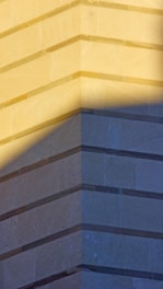 The image features a geometric structure with sharply defined lines and shadows. The corner of a building is highlighted, showing the texture and layers of the stone material. Shadows create a contrast, adding depth and dimension.