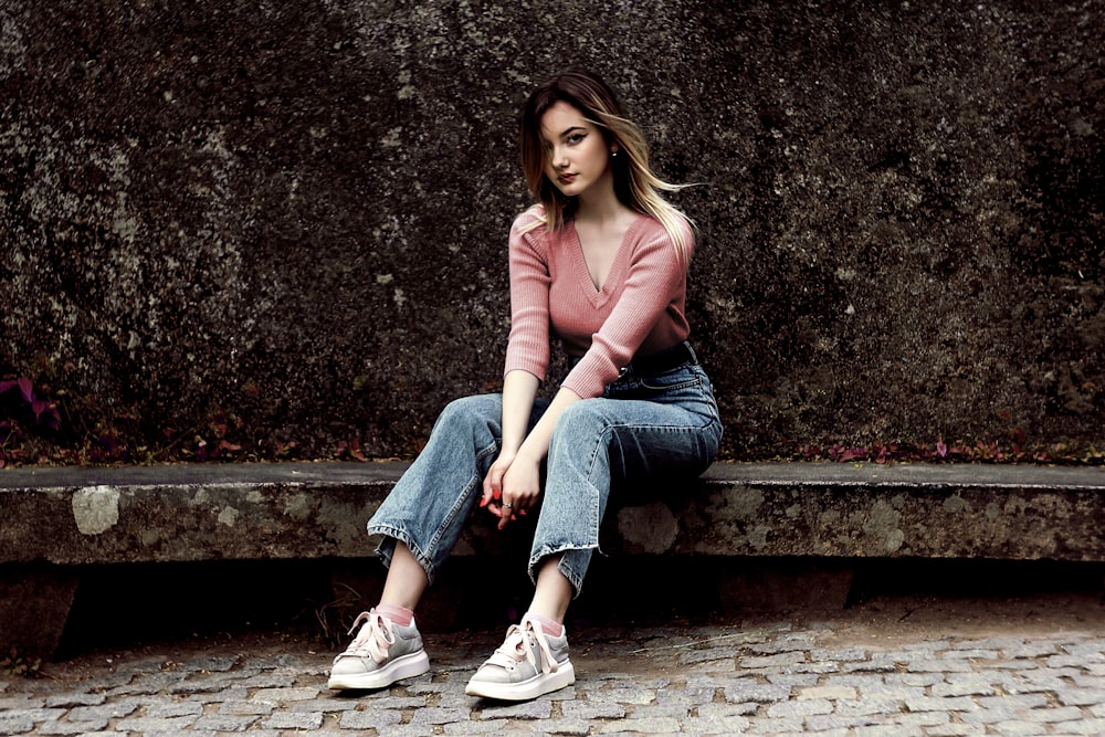 woman in pink long sleeve shirt and blue denim jeans sitting on concrete bench