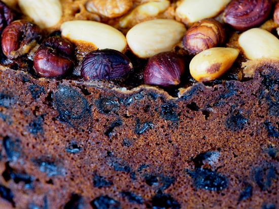 A close-up view of a fruit cake with visible nuts and dried fruits embedded on the top. The nuts include almonds and hazelnuts, while the dried fruits are dark and scattered throughout the cake. The texture appears crunchy and rich, with a deep brown color indicating a well-baked surface.
