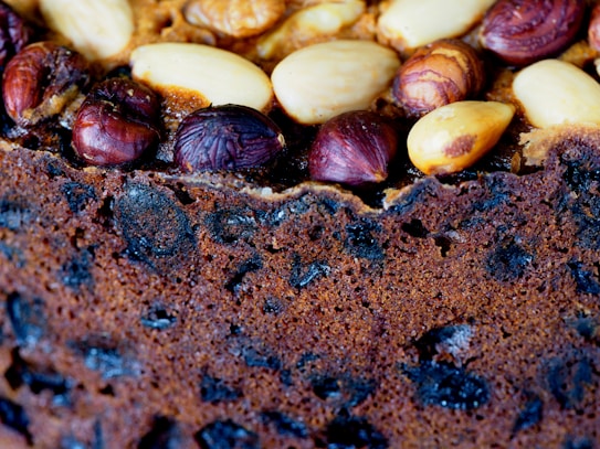 A close-up view of a fruit cake with visible nuts and dried fruits embedded on the top. The nuts include almonds and hazelnuts, while the dried fruits are dark and scattered throughout the cake. The texture appears crunchy and rich, with a deep brown color indicating a well-baked surface.