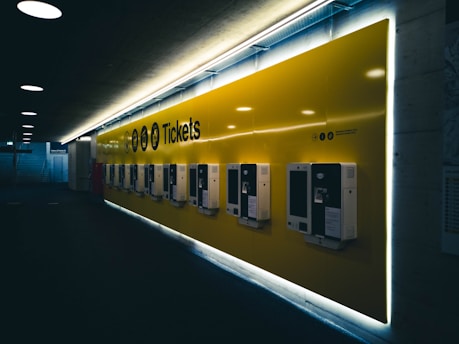 A row of ticket vending machines is mounted on a brightly lit yellow wall in a dimly lit hallway or corridor. The machines are evenly spaced and organized, and the word 'Tickets' is prominently displayed above. The lighting creates a contrast between the dark surroundings and the illuminated wall.