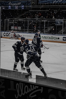 Ice hockey players in dark uniforms are engaged in a game on the ice rink, with spectators visible in the background seating area. The players appear to be focused and in motion. Advertisements and team logos are displayed around the rink.
