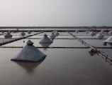 Rows of salt mounds are arranged in rectangular evaporation pools, reflecting on the water-filled surfaces. The scene appears calm with a hazy sky and a few distant figures walking along the horizon.