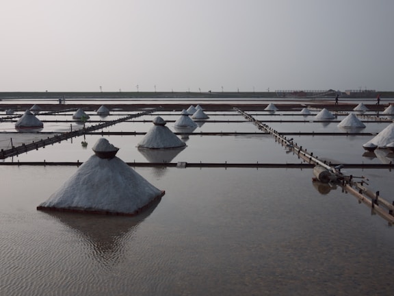 Rows of salt mounds are arranged in rectangular evaporation pools, reflecting on the water-filled surfaces. The scene appears calm with a hazy sky and a few distant figures walking along the horizon.