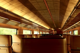 A vintage interior of a train car is depicted, featuring wooden paneling and warm lighting. The ceiling is arched and covered with an intricate pattern. Rows of seats are visible, and the windows reveal a blurred view of the passing scenery.
