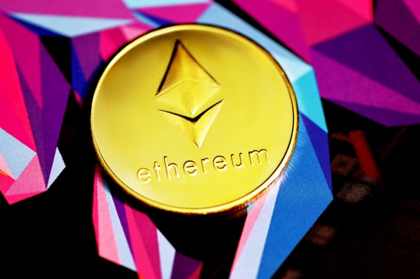 A golden coin featuring the Ethereum logo and the word 'ethereum' is placed on a vibrant background of geometric shapes in shades of purple, pink, and blue.