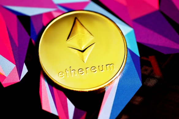 A golden coin featuring the Ethereum logo and the word 'ethereum' is placed on a vibrant background of geometric shapes in shades of purple, pink, and blue.