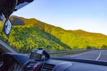 A vibrant landscape view from inside a moving car, showcasing lush green hills under a clear blue sky. The car's dashboard is partially visible, including a mounted GPS device. The road curves through the scenic countryside, bordered by trees and hills.