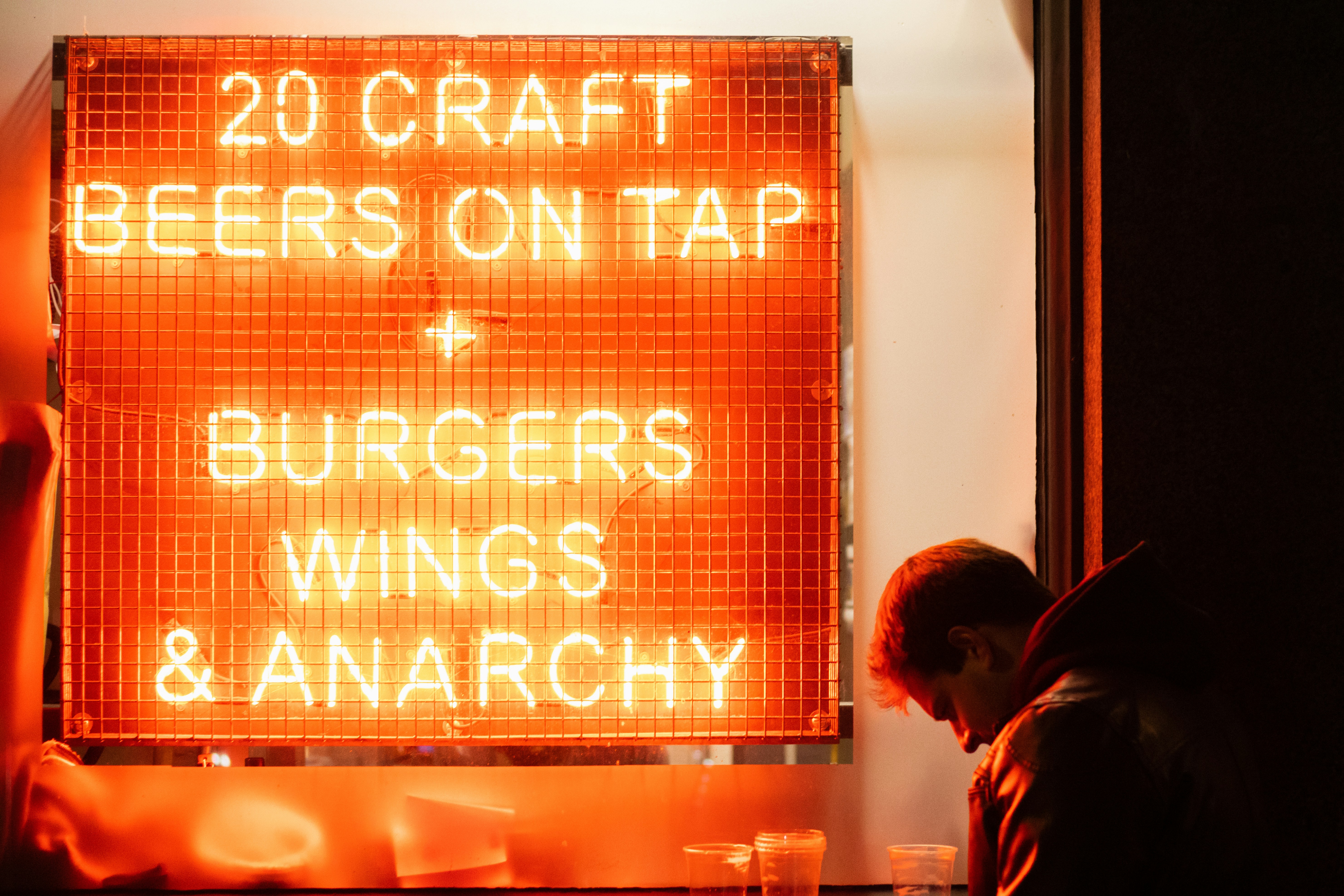A man sits, head bowed, near a large neon sign that reads "20 CRAFT BEERS ON TAP + BURGERS WINGS & ANARCHY"
