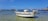 A small motorboat floats in clear, shallow water with a sandy bottom, surrounded by other boats in a serene coastal bay. The sky is clear and blue, with a few clouds scattered across it.