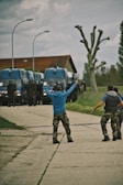 A group of individuals in casual military-style attire stand on a paved pathway, gesturing towards a line of large, blue police vans. The scene is set outdoors on a cloudy day, with a modern building and pruned trees in the background.