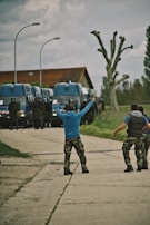 A group of individuals in casual military-style attire stand on a paved pathway, gesturing towards a line of large, blue police vans. The scene is set outdoors on a cloudy day, with a modern building and pruned trees in the background.