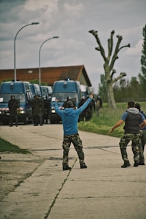 A group of individuals in casual military-style attire stand on a paved pathway, gesturing towards a line of large, blue police vans. The scene is set outdoors on a cloudy day, with a modern building and pruned trees in the background.